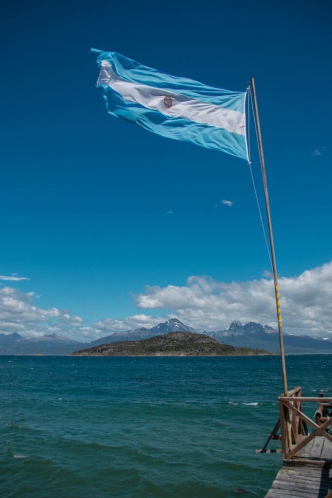 A vibrant image of the Argentine flag flying above the blue waters of Ushuaia with mountain views.